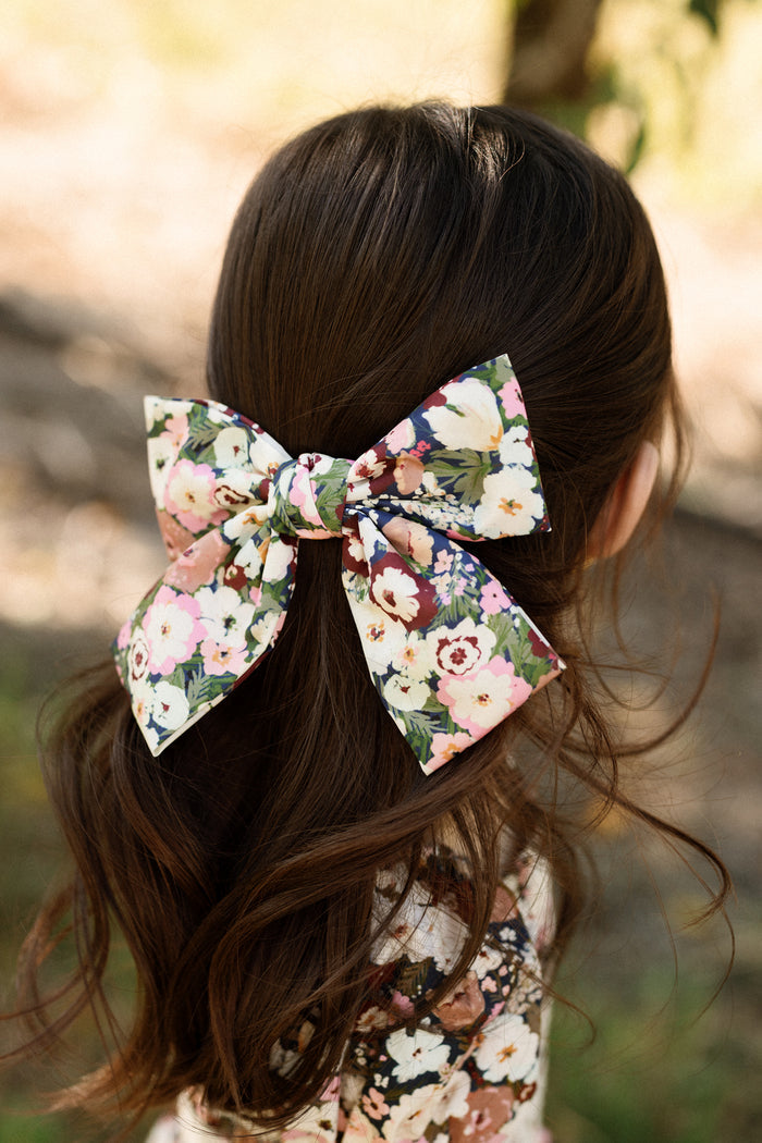 A child with long brown hair wears an Ivy City Co “Hair Bow in Midnight Garden” on the back of their head. The softly blurred background showcases natural outdoor scenery evocative of Ivy City’s enchanting Midnight Garden.