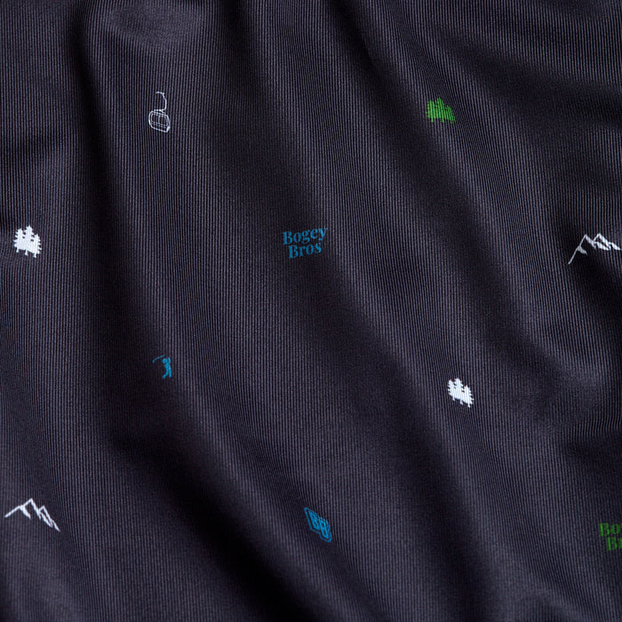 A close-up of the Pine - Polo shows dark, ribbed athletic fabric with subtle folds. Small white and green embroidered mountain, tree, and flag icons decorate it, with "Bogey Bros" in blue centered on the chest.