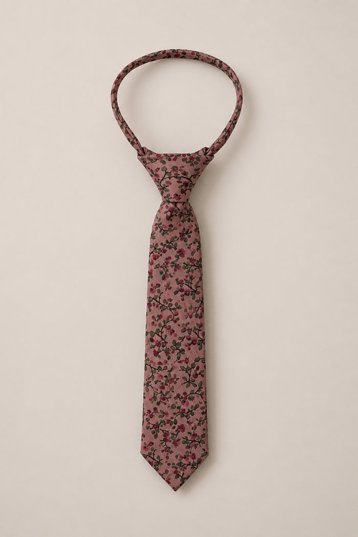 An elegant wedding accessory from Ivy City Co, the “Ties in Mauve Woodland Floral” features a pink cotton necktie adorned with a subtle floral pattern in dark red and green, arranged neatly on a plain beige background.