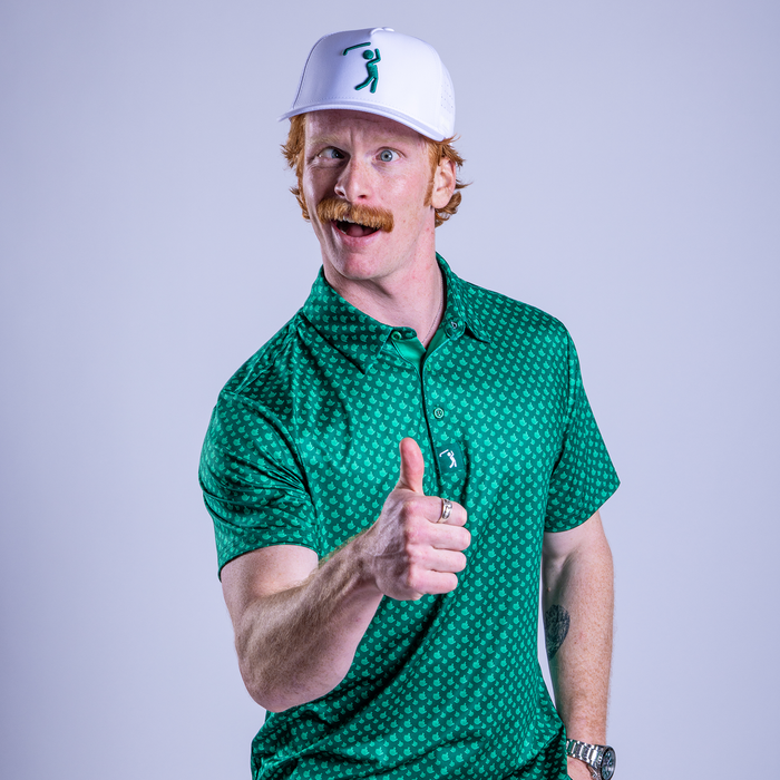 A man with red hair and a mustache, wearing a white cap and the Birdie Finger - Irish Goodbye - Polo, stands against a light purple background, smiling enthusiastically and giving a cheerful thumbs-up.