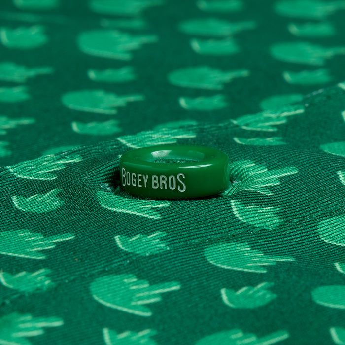 A green poker chip with "BOGEY BROS" in white rests centered on the Birdie Finger - Irish Goodbye - Polo, featuring moisture-wicking green fabric with a repeating light green pattern of raised hands and golf flags.