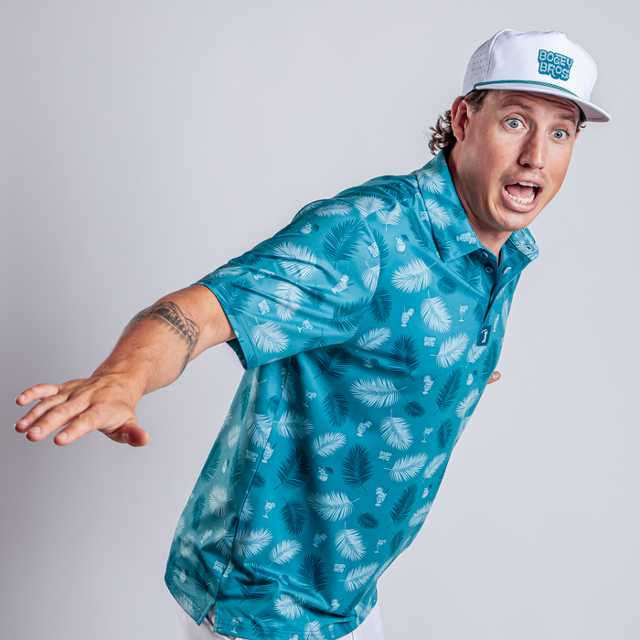 A man in an athletic fit Boat Drinks - Polo with a blue tropical print and a white “BOSS BROZ” cap leans forward, mouth open in surprise, arm outstretched, against a plain light background.