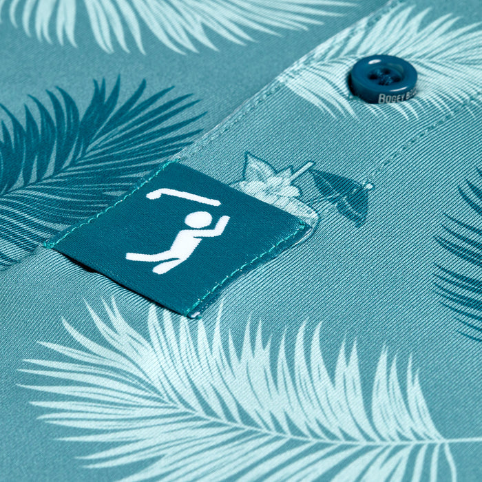 Close-up of the Boat Drinks - Polo, featuring a teal, athletic fit with a palm leaf pattern. A teal tag with a white golfer is near the button. This shirt offers moisture wicking for all-day comfort.