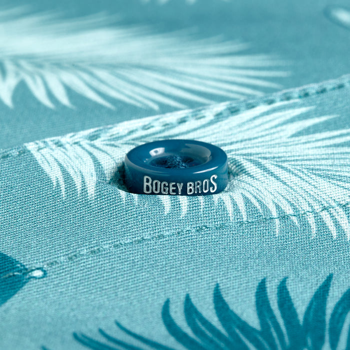 A close-up of the Boat Drinks - Polo shows a teal snap button engraved with BOGEY BROS on blue fabric with a white palm leaf pattern, offering UPF 50 protection. The button is centered, highlighting the fabric’s texture and design.