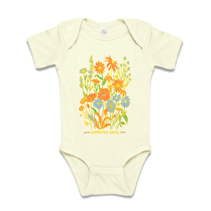 A beige baby onesie with a colorful floral pattern and the text 'Carpenter Hotel austin texas' printed on it.