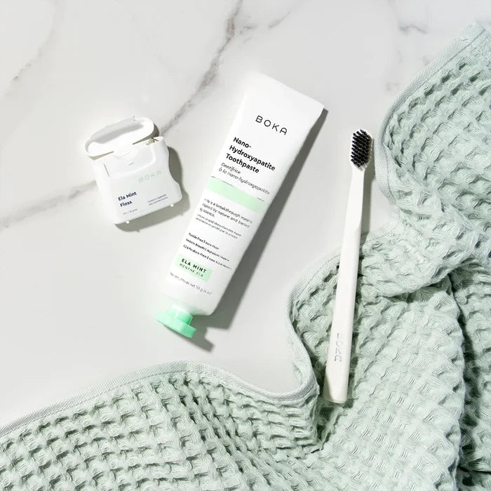 Boka toothpaste tube, toothbrush, and small container on a marble surface with a textured green towel.