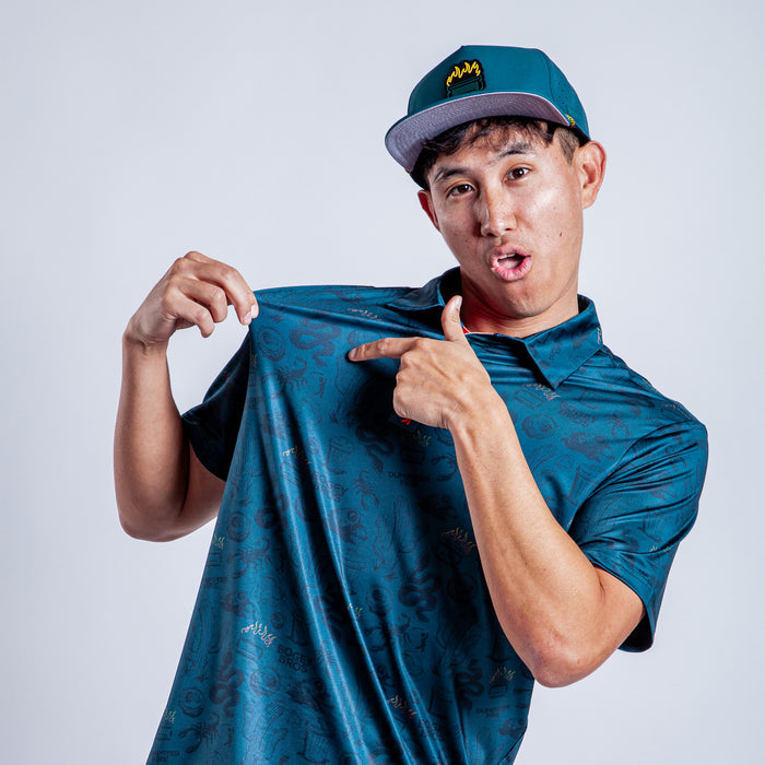 A young man in an athletic fit Deep Trouble - Polo, featuring a teal pattern, and a matching flat-brim cap playfully pulls at his shirt and points to it, smiling against a plain light gray background.
