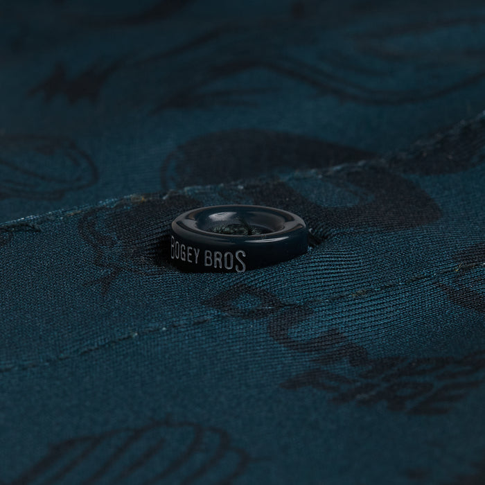 A close-up of the Deep Trouble - Polo shows a dark button on teal fabric with subtle black patterns. “BOGEY BROS” is in white on the edge. The athletic-fit polo highlights visible stitching, abstract shapes, and textured details.