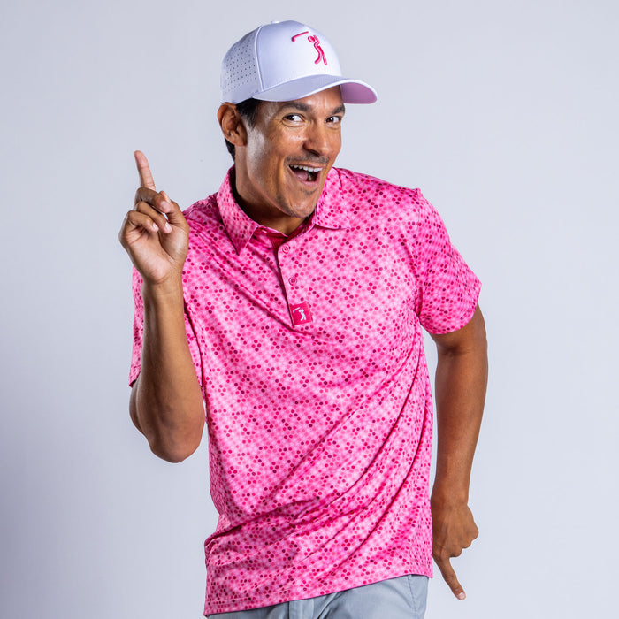 A man in the Dots - Pink - Polo, an athletic fit shirt with UPF 50 protection, smiles and points upward playfully while wearing a white and pink cap, all set against a light gray background.