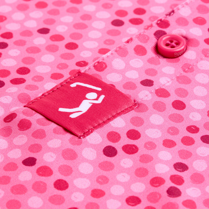 Close-up of the Dots - Pink - Polo fabric, displaying variously shaded pink dots and moisture-wicking properties. A pink button is near the top right, while a stitched patch with a white stick figure icon sits on a dark pink background at center.