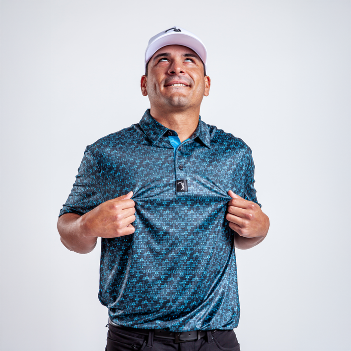 A man in a white cap and the Eyes Up Here - Zapped - Polo, featuring a blue pattern, smiles and looks upward. He stands against a white background, holding out his shirt to display the front logo and athletic fit.
