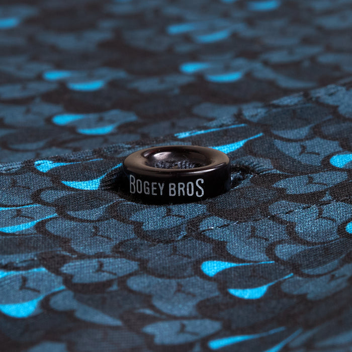 Close-up of a black metal grommet on the Eyes Up Here - Zapped - Polo, set in blue and black fabric with overlapping fish scale patterns. The centered grommet and shiny, moisture-wicking scales ensure all-day comfort.