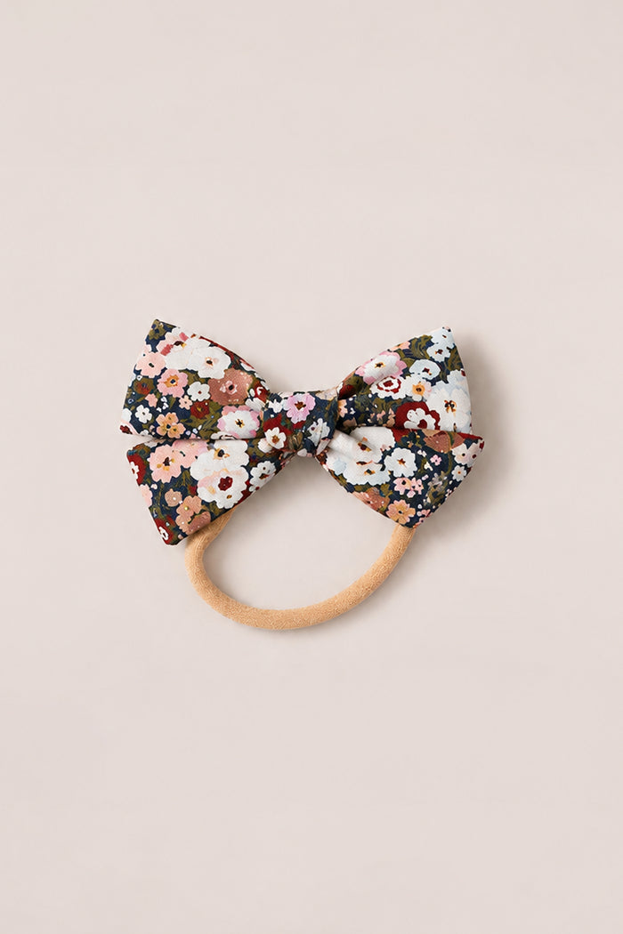 Introducing the Hair Bows in Midnight Garden by Ivy City Co—an elegant hair accessory featuring a large fabric bow adorned with a vibrant floral pattern in red, pink, white, and green. Reminiscent of Ivy City Co’s signature femininity, this charming hair tie is set on a beige elastic band and elevates any look with the enchanting spirit of Midnight Garden. Photographed delicately on a light background.