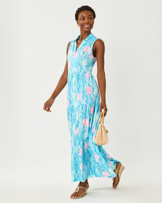 Woman wearing a blue floral maxi with teal and pink seashell details and holding a beige bag on a white background