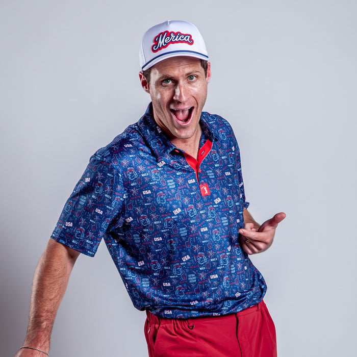 A man in a 'Merica - Navy - Polo with moisture-wicking fabric and red pants poses enthusiastically against a plain gray background, smiling with his mouth open and pointing with both hands.