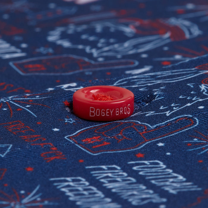 A close-up of the red BOGEY BROS button on the 'Merica - Navy - Polo, attached to blue fabric with white and red outlined prints of words like FOOTBALL, FIREWORKS, and FREEDOM. The polo features an athletic fit and moisture-wicking comfort.