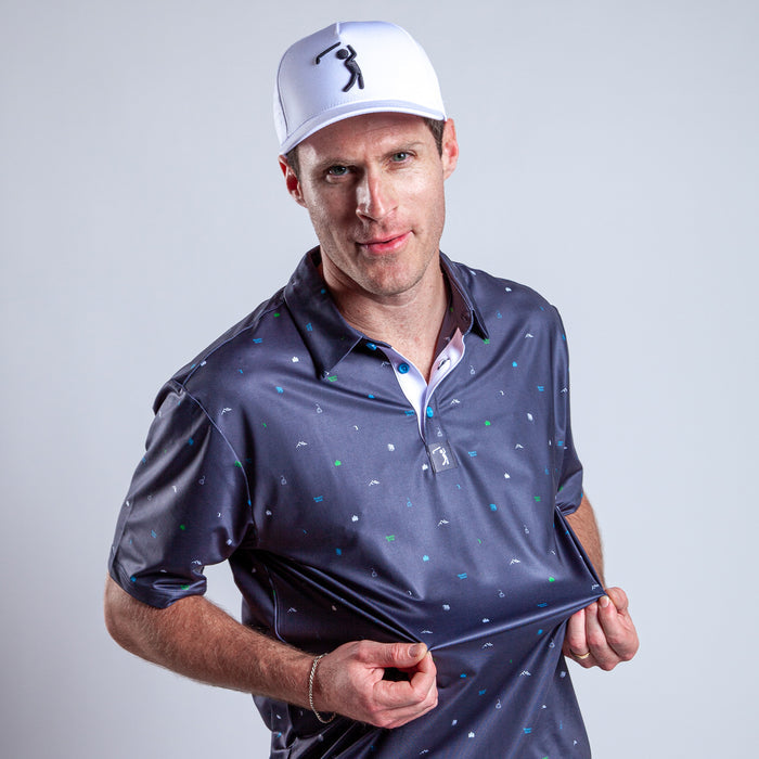 A man in a white baseball cap and navy Pine - Polo with small patterns stands against a plain light background, slightly smiling as he tugs at the bottom of his athletic fit, moisture-wicking shirt with his right hand.