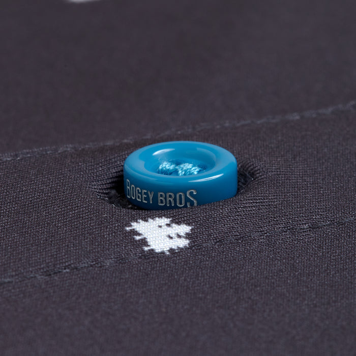 Close-up of a blue button sewn onto dark fabric on the Pine - Polo, with BOGEY BROS printed around the edge. The moisture-wicking, athletic-fit fabric shows white pixel-style graphics near the button, which is secured with blue thread.
