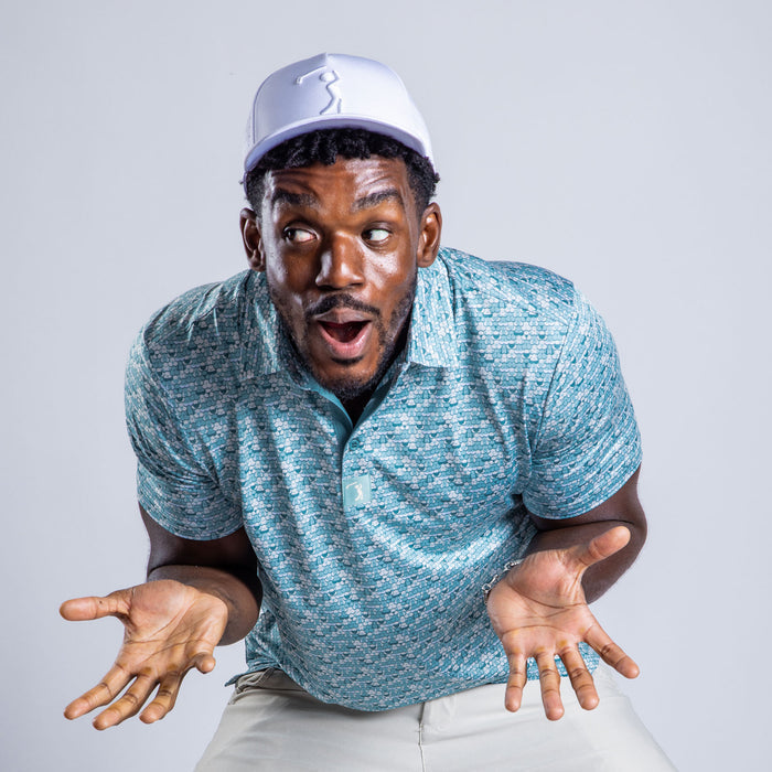 A man wearing the T&A - Luscious - Polo, a moisture-wicking patterned teal shirt, and a white cap looks surprised with wide eyes and mouth open, leaning forward with hands out against a plain light background.
