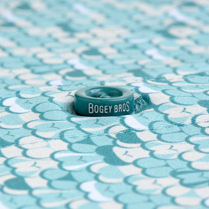 A teal “BOGEY BROS” button is sewn onto the T&A - Luscious - Polo, featuring moisture-wicking fabric with overlapping teal, mint, and cream scallop patterns. The button is centered in a small dimple on the shirt.