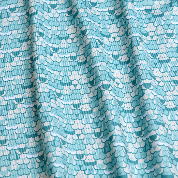 A close-up of the T&A - Luscious - Polo, featuring light blue moisture-wicking fabric with a turquoise and white repeating scallop pattern. Soft folds create gentle shadows, making it perfect for staying cool and dry.
