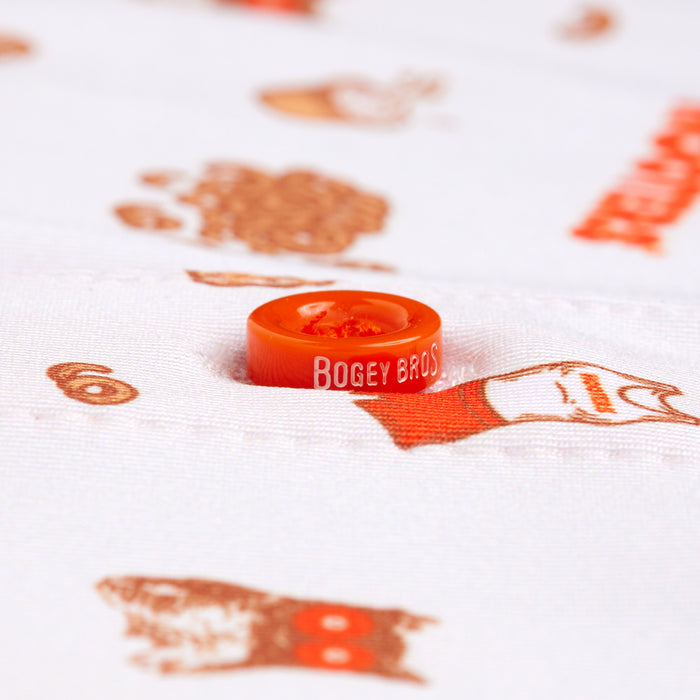 A close-up of an orange button embossed with BOGEY BROS. on the Wingin' It - White - Polo, featuring white moisture-wicking fabric with small brown and orange cartoon faces and objects, plus part of an orange word on the right side.