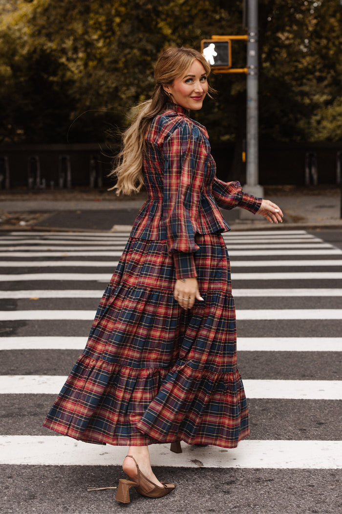 Wearing Ivy City Co’s Zoey Skirt in Twilight Plaid and chic heels, a woman strolls across a crosswalk, smiling over her shoulder—a quintessential fall fashion moment. Autumn trees and an illuminated pedestrian signal set the scene behind her.