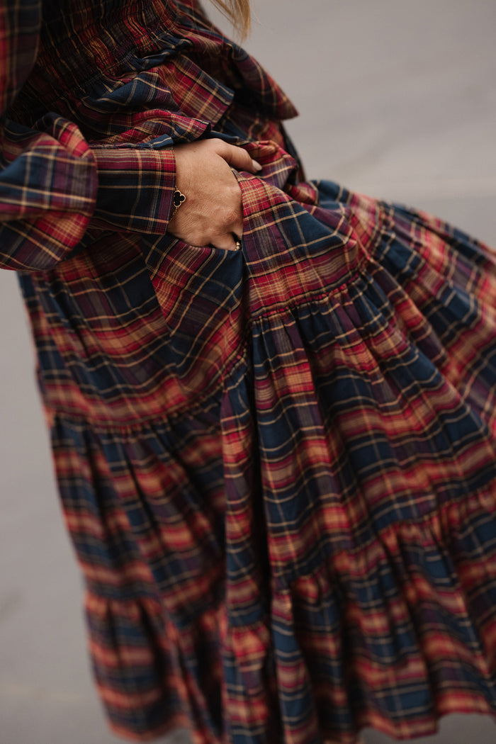 A person wearing the Zoey Skirt in Twilight Plaid by Ivy City Co holds a handful of the soft cotton A-line skirt, creating gentle folds in its red and navy plaid fabric. The neutral, out-of-focus background highlights an effortless fall fashion moment.
